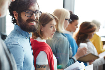 Cheerful excited handsome bearded man in denim shirt wearing eyeglasses standing in line and...