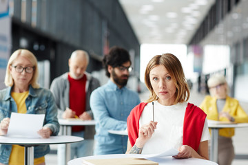 Serious thoughtful confident attractive lady filling application form and thinking about answers while applying it to register in event, she standing at high table in lobby