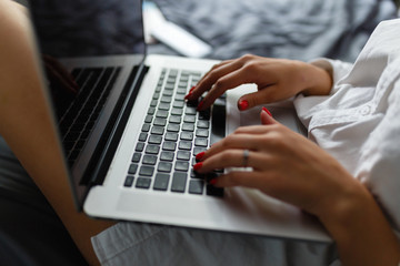 Hands close-up of a young girl in a man's shirt lying on the bed in the bedroom with a laptop on her lap and working. Work at home. The girl checks the mail in the morning on the tucked bed