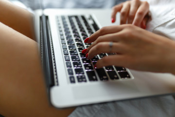 Hands close-up of a young girl in a man's shirt lying on the bed in the bedroom with a laptop on her lap and working. Work at home. The girl checks the mail in the morning on the tucked bed