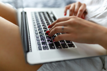 Hands close-up of a young girl in a man's shirt lying on the bed in the bedroom with a laptop on her lap and working. Work at home. The girl checks the mail in the morning on the tucked bed