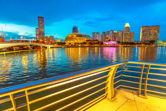 Skyline of marina bay with a bridge with evening lights reflecting in the sea at blue hour. Singapore cityscape by night. Night scene waterfront promenade.