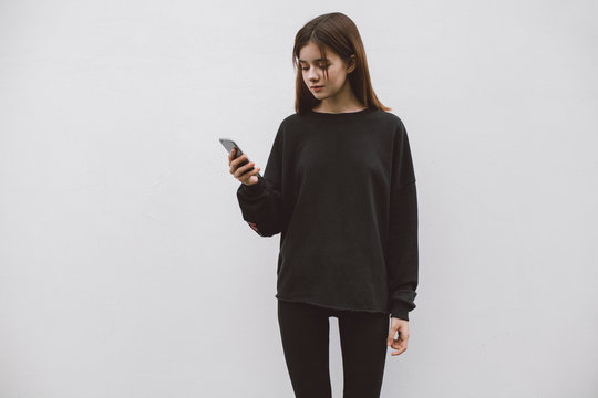 Hipster female holding in her hand a modern smartphone standing on the background of a white wall. Blank hoodie with empty place for logo, text or design. Black blank sweatshirt mockup