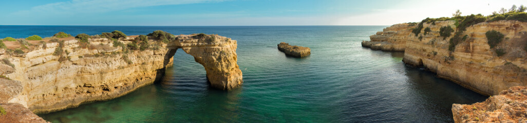 Panoramic view of the natural arch Arco  da Albandeira in the Algarve, Portugal. The arch and surrounding cliffs as well as a rock stump in the Atlantic ocean are shown on a sunny day with calm water.