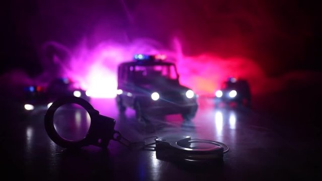Police raid at night and you are under arrest concept. Silhouette of  handcuffs with police car on backside with the flashing red and blue police lights at foggy background. Slider shot