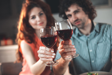 Cheers. Happy loving couple clinking glasses of wine while sitting in kitchen. Focus on red wine 