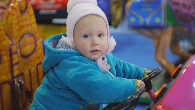 Portrait of 1.5 years old blue eyed babe playing coin operated kiddie ride. Baby girl turns steering wheel of a toy car