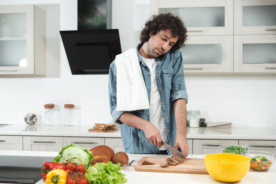 Portrait of cheerful young man is cutting bread in kitchen while talking on smartphone. He is standing at table while carrying a towel on his shoulder 