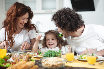 Happy childhood. Cheerful little girl is laughing while her father is keeping piece of broccoli near her. Mother is looking at them with love and smiling while eating salad 