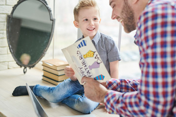 Lovely moments of fatherhood: joyful bearded man reading handmade greeting card given by his little son for Fathers Day while sitting at modern living room