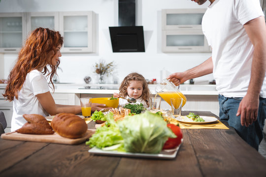 Caring mom and dad are feeding their child with healthy breakfast. Woman is holding a bowl with salad while man is pouring juice into glass. Pretty girl is looking at food with appetite 