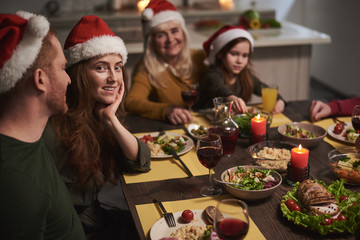 Portrait of smiling female sitting next to her husband at festive dinner. They are wearing red hats and eating wholesome dishes with red wine