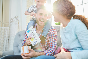 Mischievous little boy sitting on shoulders of his bearded dad and pulling his ears while he receiving gifts and greeting cards for Fathers Day from family members, lens flare