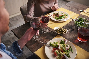 Dinner for two. Close up of couple hands clinking glasses. Female and male are sitting at table eating salads and vegetables and drinking red wine