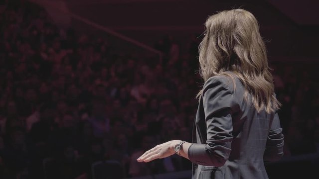 The female financial coach emotional gesturing talks from the stage with spectators at forum. Too many anonymous persons workers and students seat in large auditorium and watch workshop background