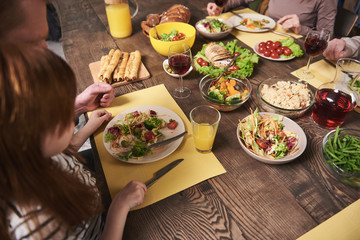 Close up of wooden table full of wholesome meal including salads, vegetables, meat and wine. Family members are sharing food together in kitchen at home