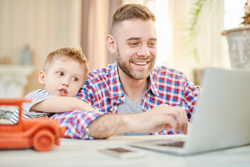 Fototapeta na wymiar Cheerful bearded man wearing checked shirt and his cute little son playing computer game together while spending weekend at home