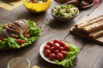 Close up of baked meat lying on lettuce together with white plate of cherry tomatoes. Bread and salads in bowls are nearby