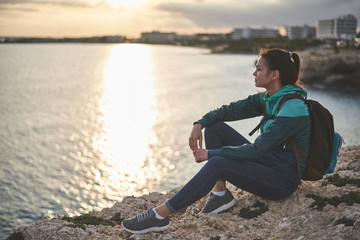 Calm girl is watching sunset at the seashore. She is sitting on stone with beautiful landscape on background 