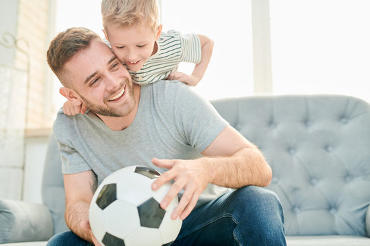 Happy young father and his cute little son having fun together while taking break from playing football, interior of cozy living room on background
