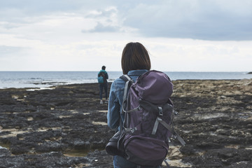 Back view of young woman hiking on rocky seashore. She is carrying backpack. Her friends is going on background 