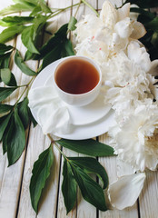 White porcelain cup of morning tea and white fresh peonies on light gray wooden background. Top view, concept of marriage proposal.