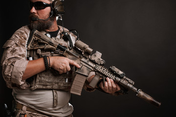 Brutal man in the military desert uniform and body armor stands in a fighting rack and holds his...