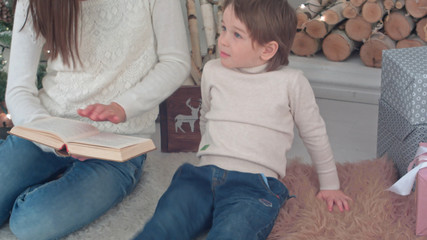 Capricious boy listening to his mom reading a book near Christmas tree