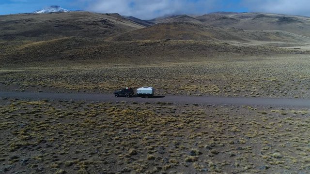 Aerial drone scene of van and trailer, motorhome in steppe, patagonia argentina riding on a gravel lonely road. Tromen volcano on the background. National Park. Camera moving left tracking car.
