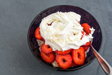 Cooked oatmeal in a purple glass bowl with fresh sliced strawberries and whipped cream on top and a spoon in the bowl, on a slate background
