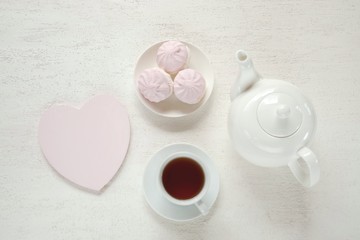 Morning tea flat lay. White cup with tea, white teapot, pink marshmallow on a light relief background.top view, copy space