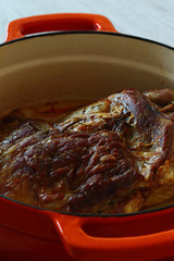 Roasted shoulder of lamb in an orange ceramic roasting dish after cooking on slate and grey wood kitchen worktop
