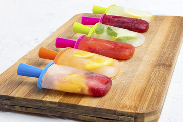 Homemade colorful fruity popsicle on a wooden board and against a white stone background. Strawberry, Lemon, Lemon with mint, Orange, Cherry, Multifruit