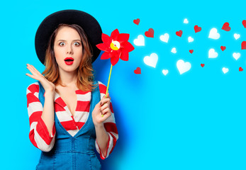 Fototapeta na wymiar Portrait of young surprised red-haired white european woman in hat and red striped shirt with jeans dress with pinwheel on blue background with hearts