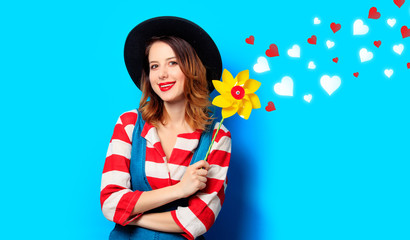 Portrait of young smiling red-haired white european woman in hat and red striped shirt with jeans dress with pinwheel on blue background with hearts