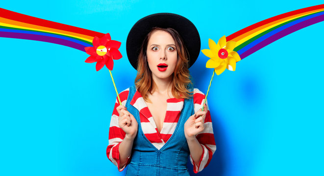 Portrait of young surprised red-haired white european woman in hat and red striped shirt with jeans dress with pinwheel on blue background and with rainbow