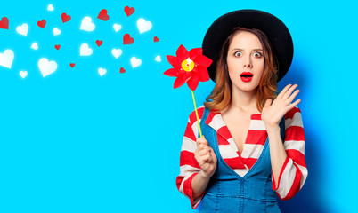 Portrait of young surprised red-haired white european woman in hat and red striped shirt with jeans dress with pinwheel on blue background with hearts