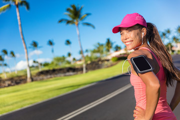 Fitness motivation running music woman runner. Happy jogging girl listening to smartphone app with earphones. Asian athlete wearing cap and armband smart phone holder outdoor on city road.