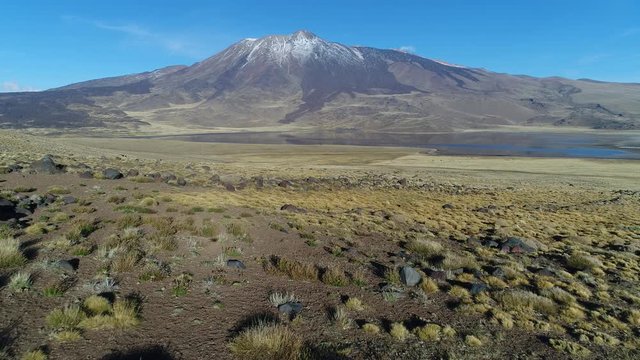 Tromen volcano, national park, Patagonia. Aerial drone scene moving forwards to the mountain with bed of lava and snow. Tromen lagoon with refletion in front of volcano. Emtpy gravel road