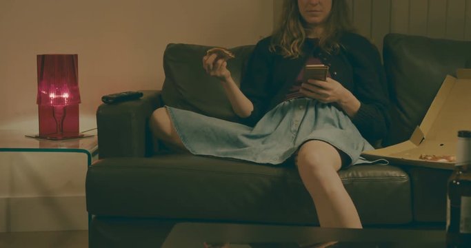 Young woman at home on sofa eating pizza and using smartphone