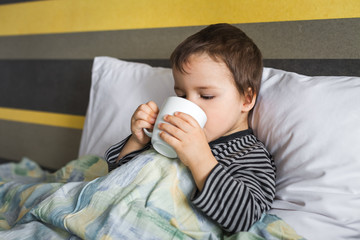 sad ill curing boy sitting on bed on pillow under blanket drinking medicine in cup