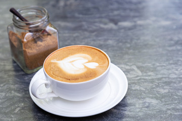 Cup of hot coffee with heart pattern in white cup and brown sugar on stone table background.