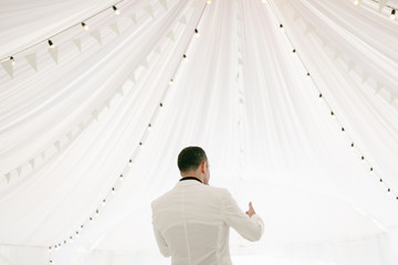 Young man in white jacket standing on the background of white tent with lanterns garland. Show...