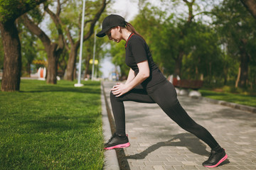 Young athletic beautiful brunette girl in black uniform, cap doing sport stretching exercises, warm-up before running or training, standing in city park outdoors. Fitness, healthy lifestyle concept.