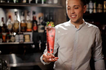 Smiling bartender holding a transparent red cocktail in the glass on the bar counter
