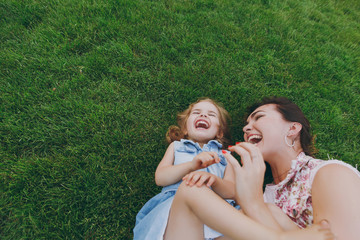 Overjoyed woman in light dress and little cute child baby girl lie on green grass in park rest play and have fun. Mother, little kid daughter. Mother's Day, love family, parenthood, childhood concept.