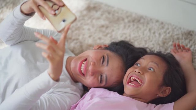 Happy young mother and her daughter taking selfie photo together with a mobile phone while lying on the carpet at home. Shot in 4k resolution