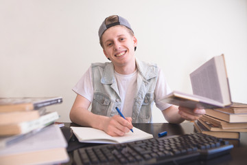 Portrait of a happy student sitting at the table with a book in his hands looking at the camera and smiles. A teenager with a pen and book in his hand makes a homework at home in the room