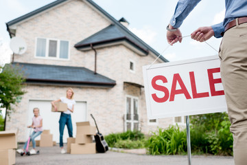 Cropped image of male realtor with sale signboard and family with cardboard boxes behind