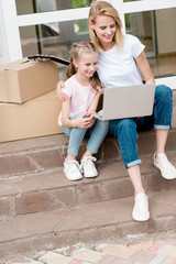 happy woman with daughter using laptop on stairs with cardboard boxes and guitar in front of new house